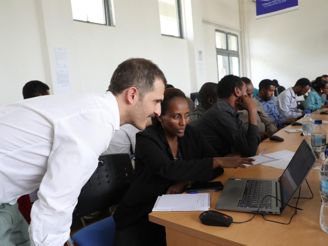 A man is bent over a laptop of his colleague to whom he is explaining something.