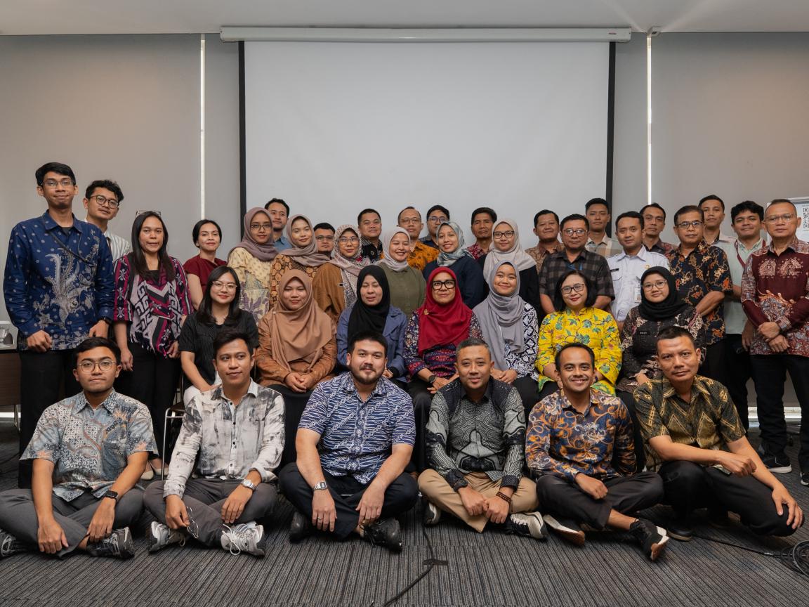 Group photograph of approximately 35 people posing indoors in front of a blank projection screen and whiteboard. Participants wear a mix of casual and traditional attire, with some seated on the floor and others standing behind.