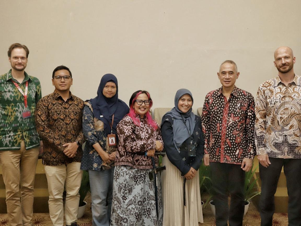 Photograph of seven people, three women and four men, standing indoors and wearing traditional patterned clothing in various colors, including green, brown, blue, and white.