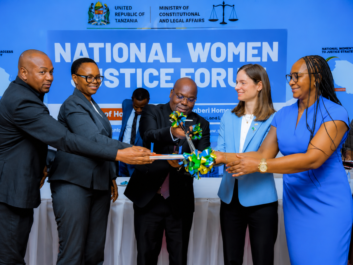 A group of officials and guests participate in a ceremonial ribbon-cutting during the launch of the National Women Justice Forum in Tanzania. The event takes place on a stage with a large backdrop displaying the forum's name alongside the logos of the United Republic of Tanzania and the Ministry of Constitutional and Legal Affairs. Six people stand at the front, smiling as they jointly cut a green, yellow, and blue ribbon tied around a publication or plaque.