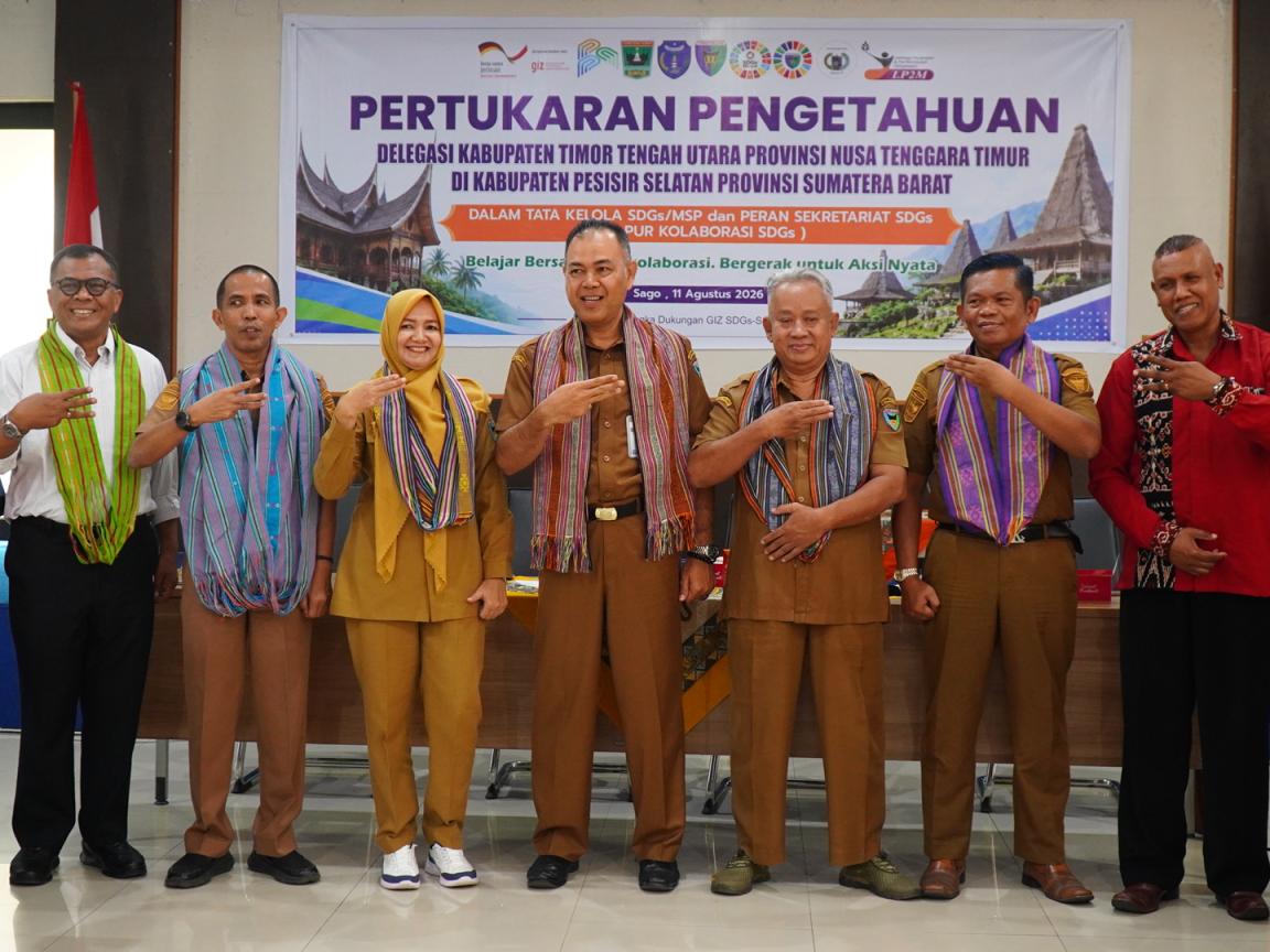 A group of nine people standing in a row indoors, posing for a formal event with a banner behind them titled "Pertukaran Pengetahuan". Participants wear traditional woven scarves and formal attire, with some giving three-fingers gestures, highlighting cultural collaboration and official partnership.