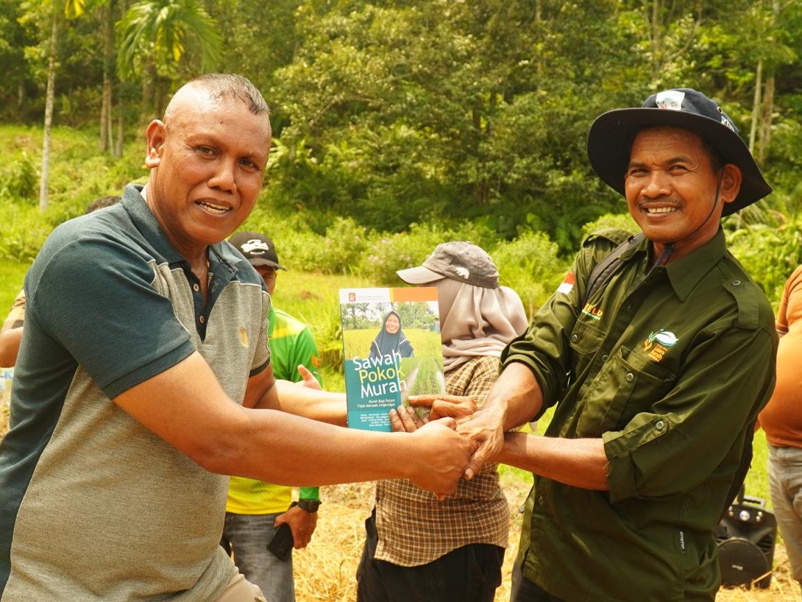 Photograph of two men outdoors shaking hands and holding a book titled "Sawah Pokok Murah" in a rural setting with greenery and other people in the background. The image captures a moment of exchange or presentation, highlighting camaraderie and community engagement.