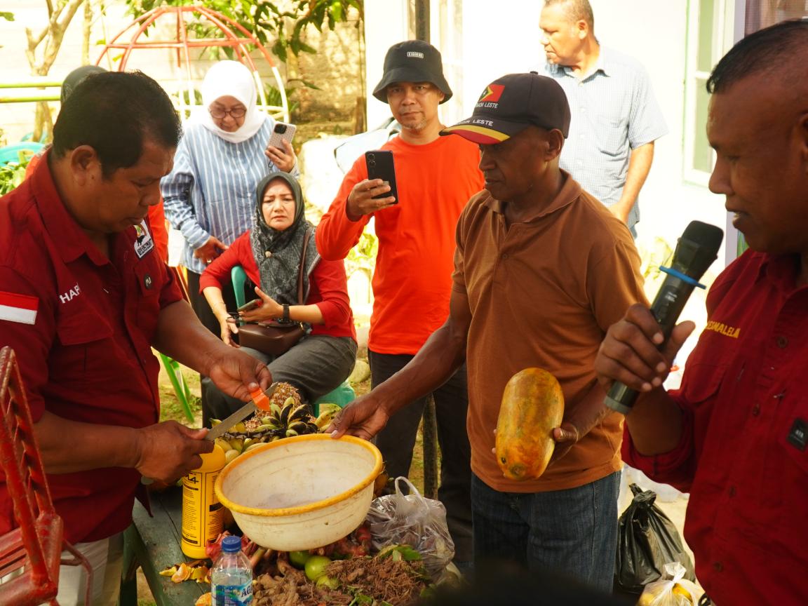 Photograph showing a group of people gathered around a table where a man in a red shirt is preparing fruit using a tool. One man holds a microphone and another holds a yellow fruit, while others observe and record the activity, suggesting a demonstration or educational event.