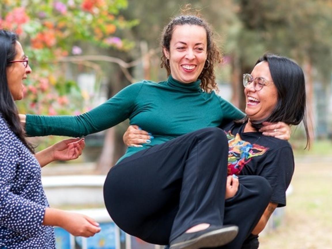 Drei Frauen stehen in einem Kreis beisammen in einem Park. Die Frau rechts hat eine andere Frau auf dem Arm.