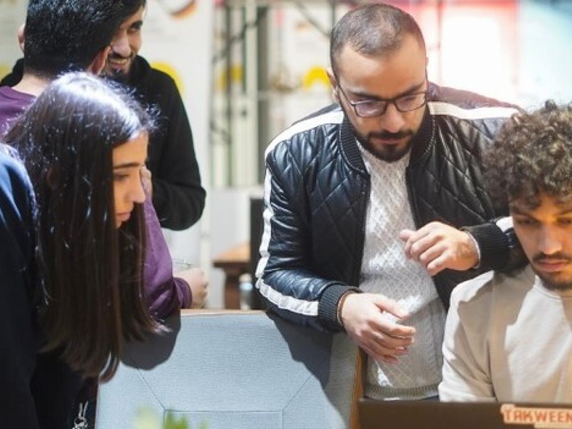 Fünf junge Menschen in einem modernen Raum, drei davon blicken auf ein Laptop, zwei unterhalten sich.