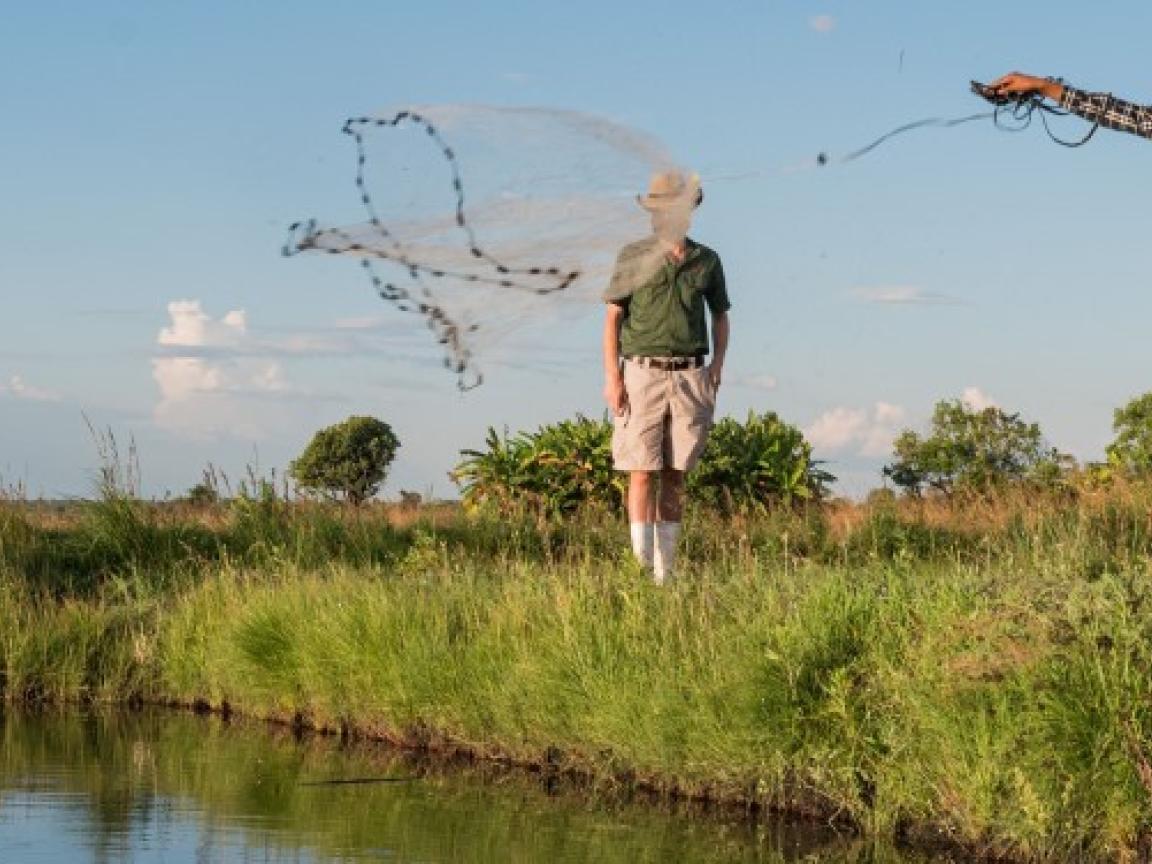 Ein Mann steht an einem Teich und wirft ein Fischernetz aus, im Hintergrund steht ein zweiter Mann.
