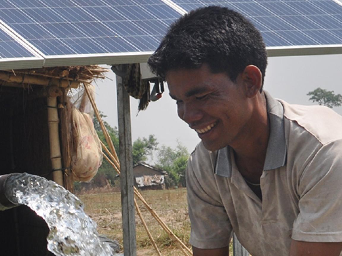 Ein Wasserschwall aus einem Rohr und ein lächelnder Mann vor einer Hütte mit Solarzellen auf dem Dach.