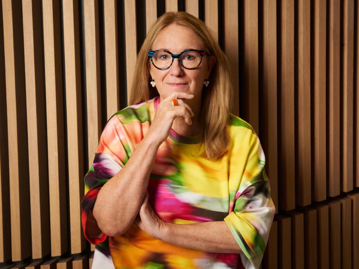 Eine Frau mit langen blonden Haaren und Brille steht vor einer Wand mit Holzlamellen und trägt eine bunte Bluse.