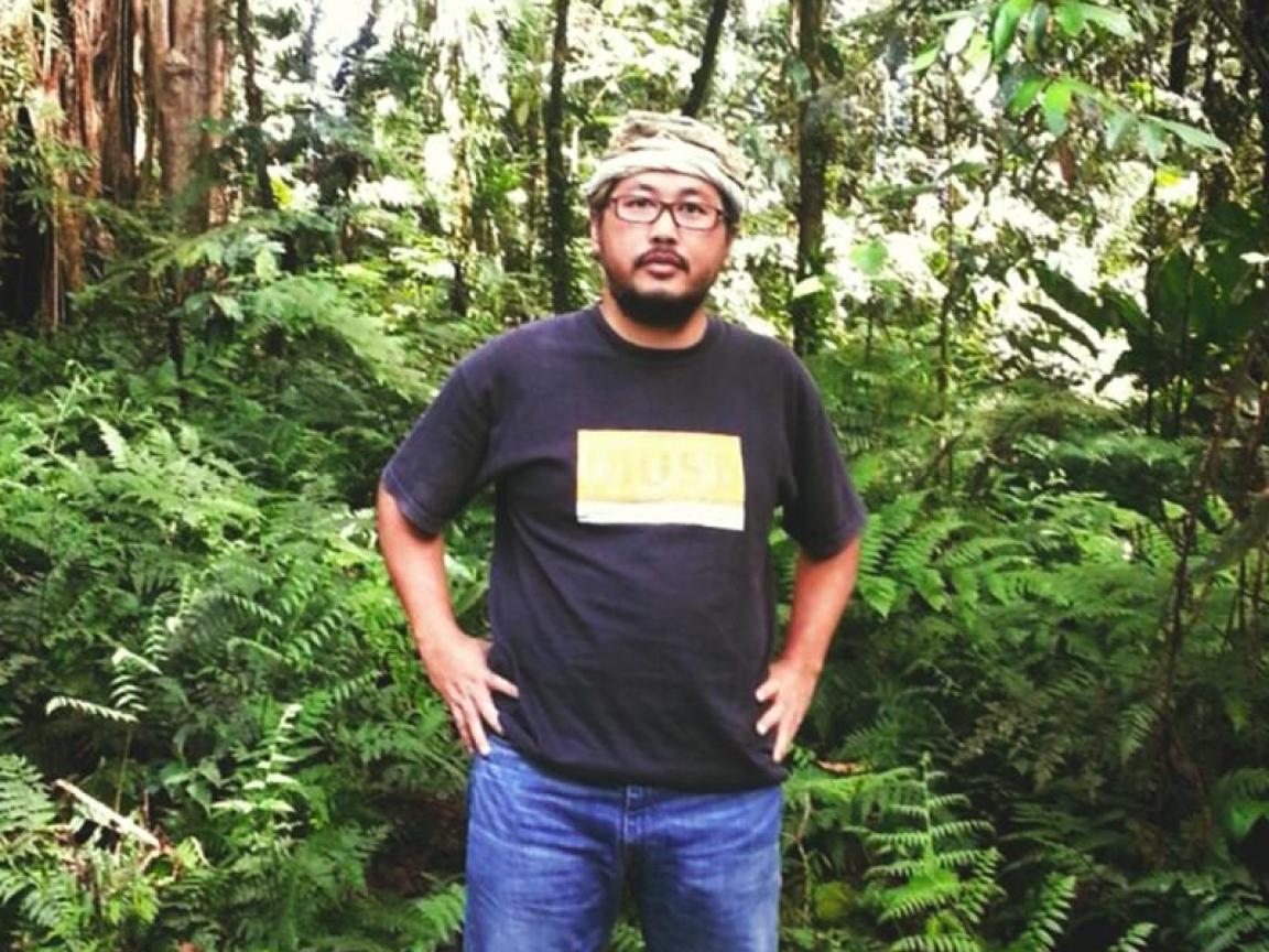 Ein Mann mit Brille, schwarzem T-Shirt und Jeans steht inmitten eines dichten, tropischen Regenwaldes mit üppigem Farnbewuchs und hohen Bäumen.