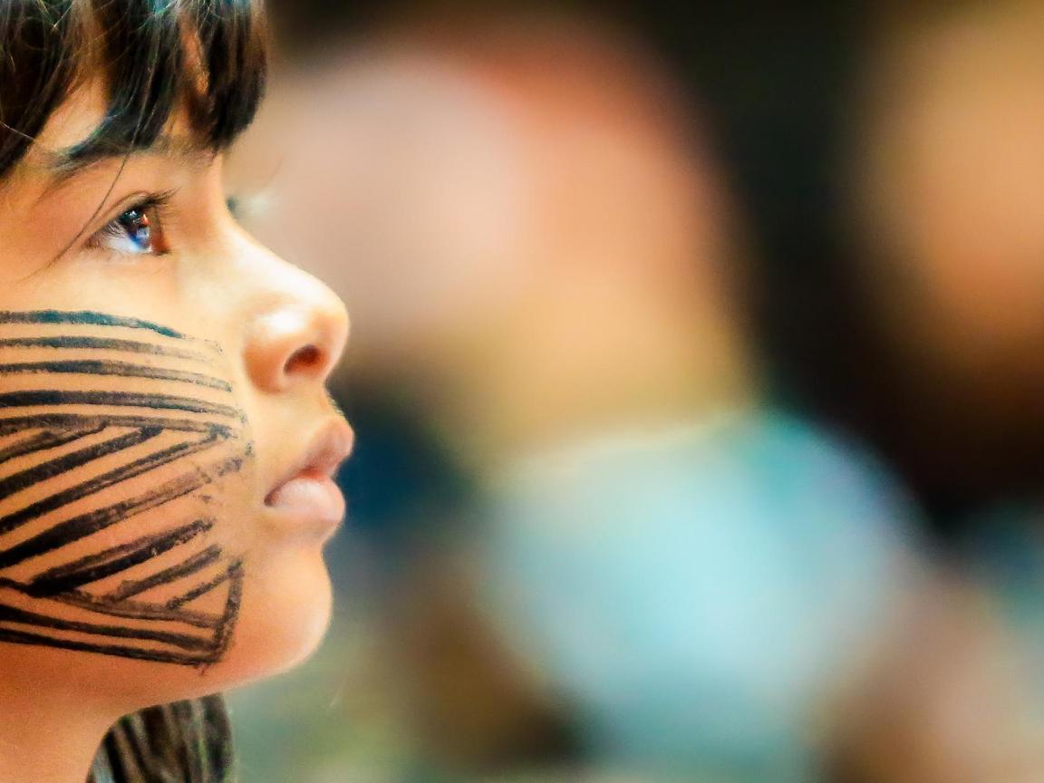 Nahaufnahme eines Kindes mit schwarzen Streifenmustern im Gesicht, das zur Seite blickt, mit verschwommenem Hintergrund.