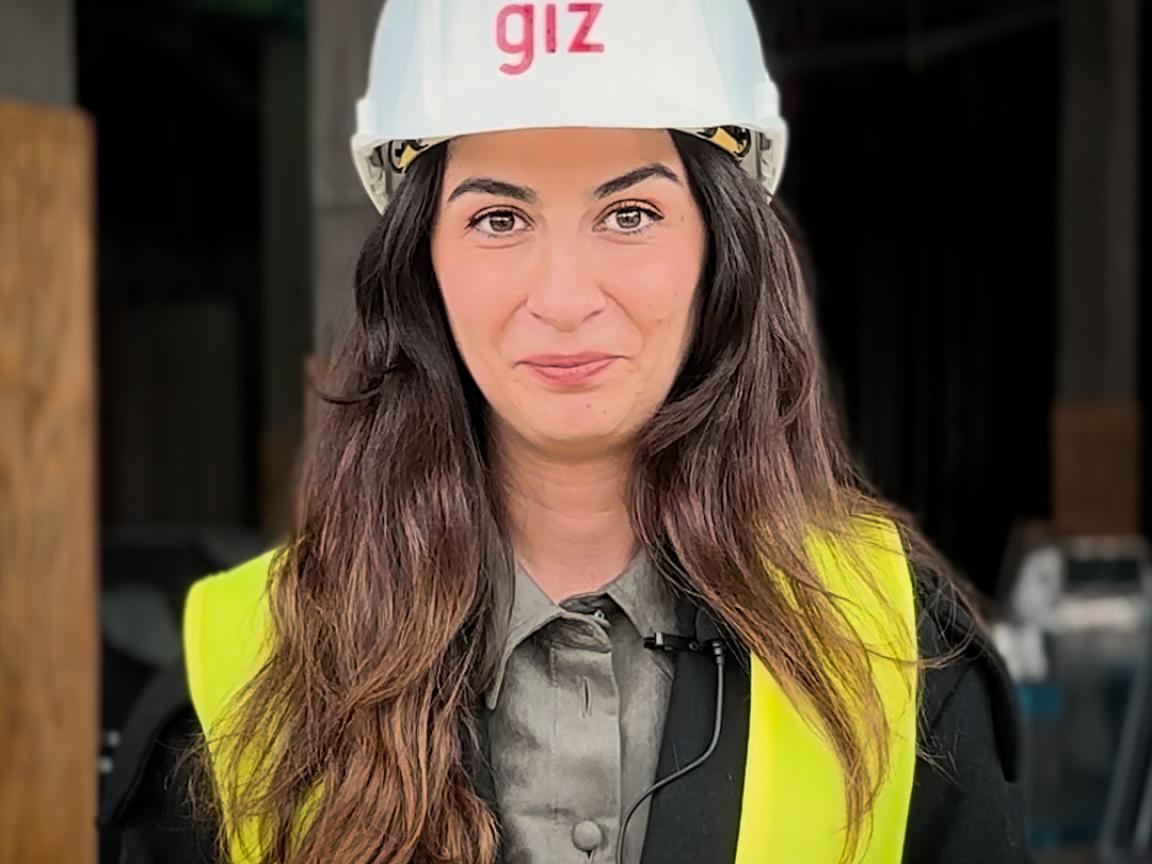 Eine Frau mit langen braunen Haaren trägt einen weißen Schutzhelm mit der Aufschrift "giz" und eine gelbe Warnweste auf einer Baustelle.