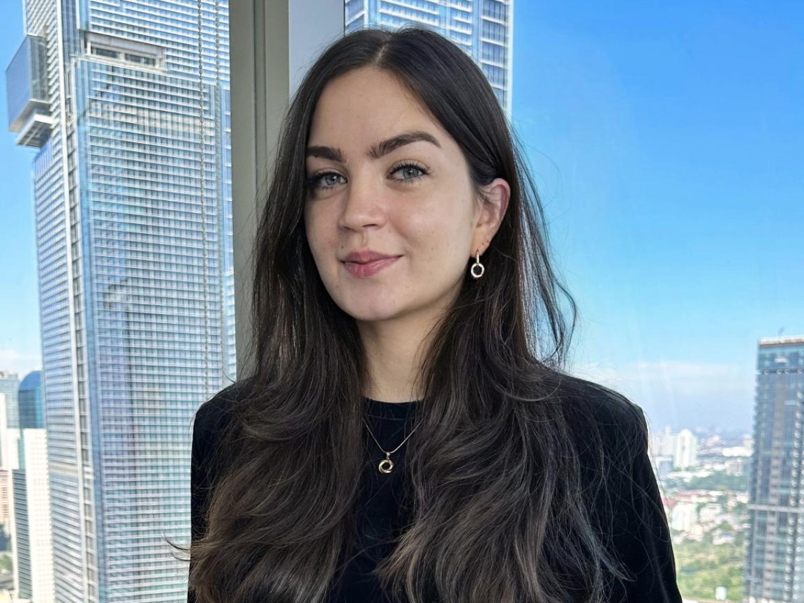 Eine Frau mit langen, braunen Haaren und schwarzem Oberteil steht vor einer Fensterfront mit Blick auf moderne Hochhäuser und eine Stadtlandschaft.