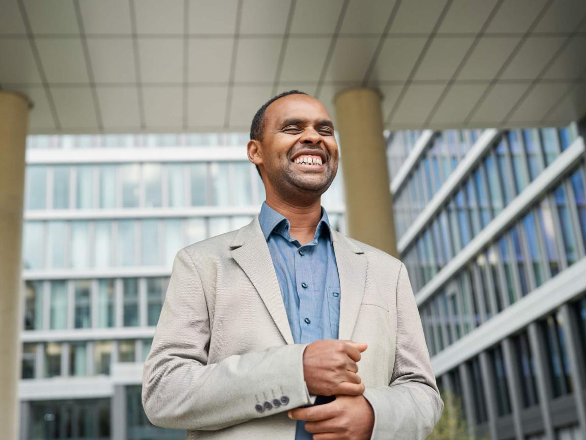 Ein lächelnder Mann in einem hellen Sakko und blauen Hemd steht vor einem modernen Bürogebäude mit Glasfassade.