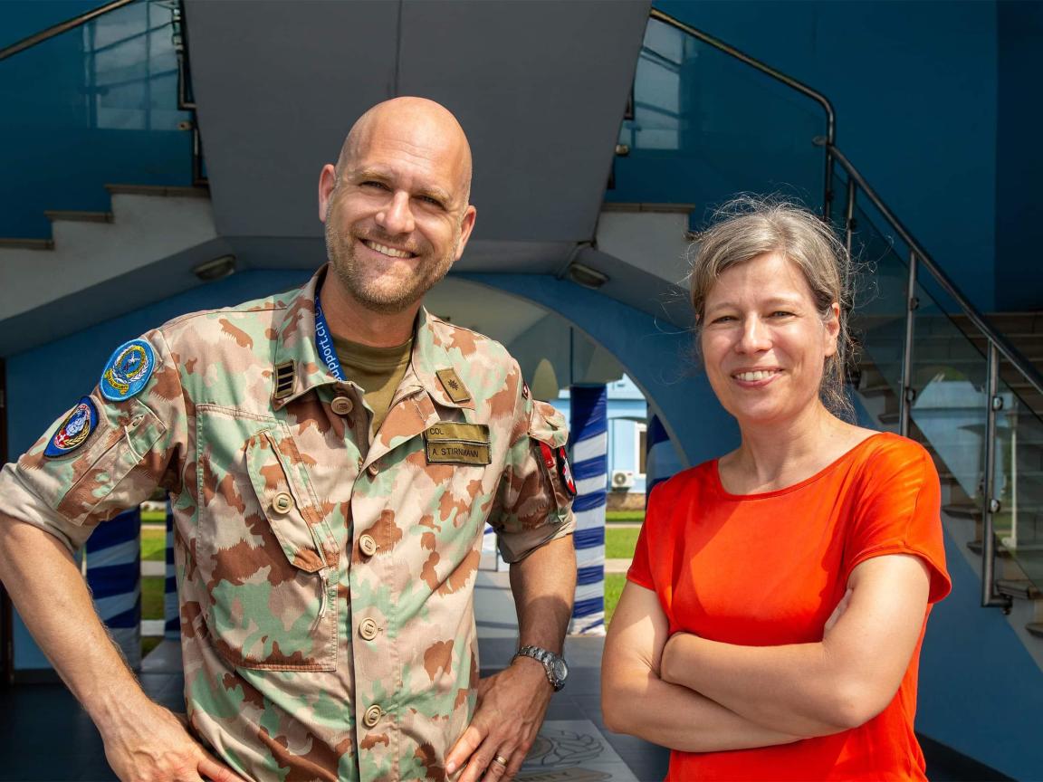 Ein lachender Mann in einer militärischen Tarnuniform mit Namensschild „A. Stirnimann“ steht neben einer Frau in einem roten Shirt vor einer modernen blauen Gebäudestruktur.