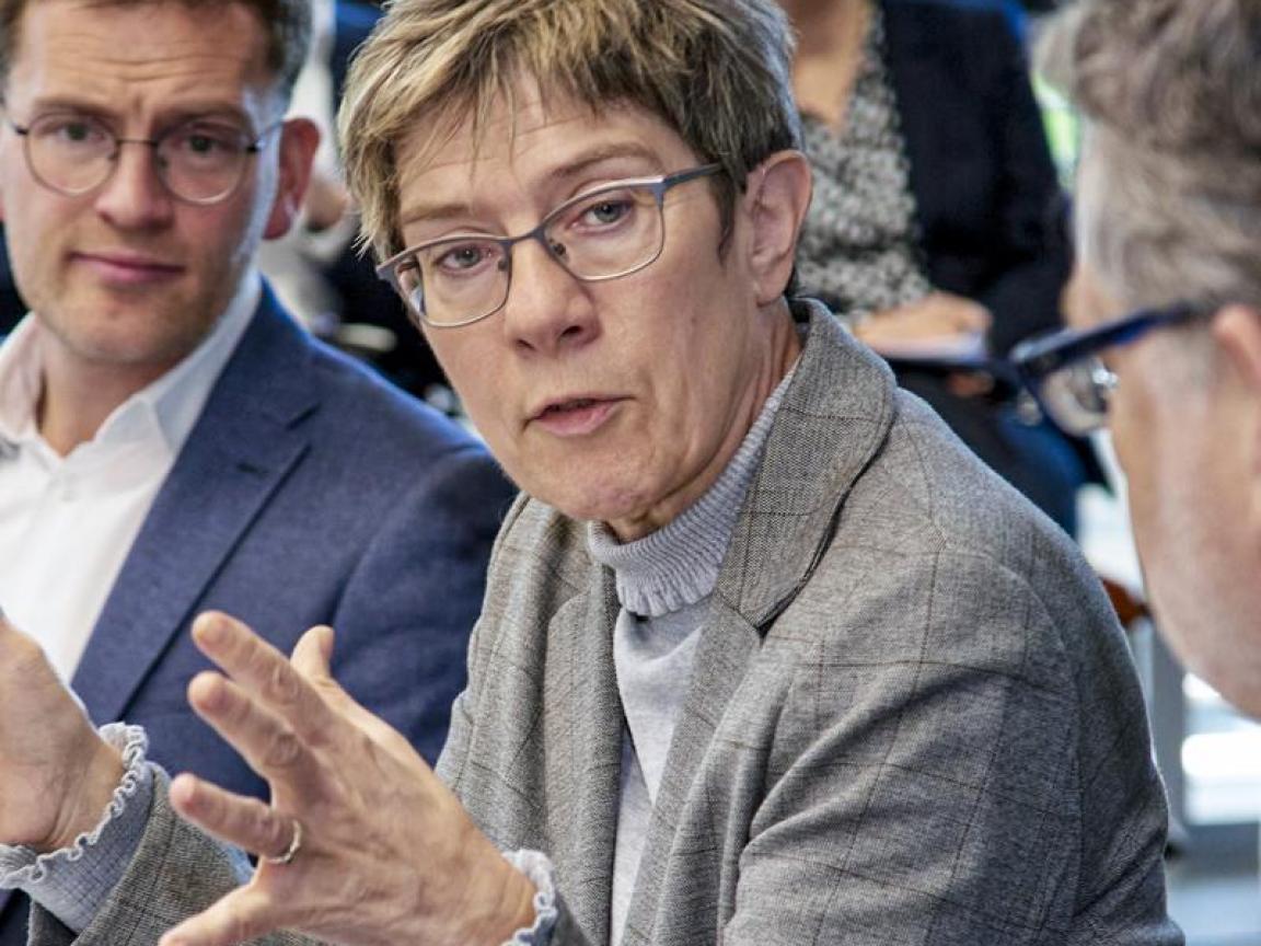 Diskussion in einem Konferenzraum mit einer Frau im grauen Blazer, die mit Gestik spricht, umgeben von mehreren Personen in Businesskleidung, die aufmerksam zuhören.