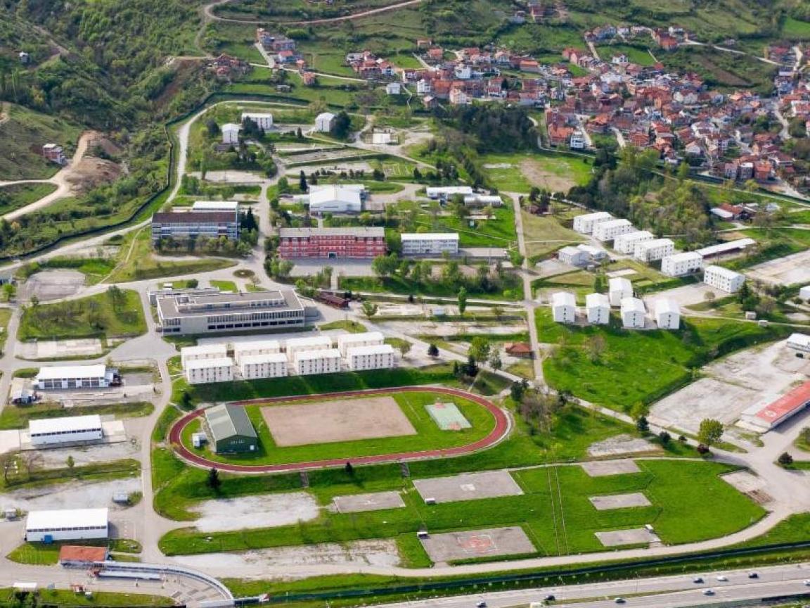 Luftaufnahme eines weitläufigen Geländes mit Gebäuden, Sportanlagen und einer angrenzenden Stadt in einer hügeligen Landschaft.