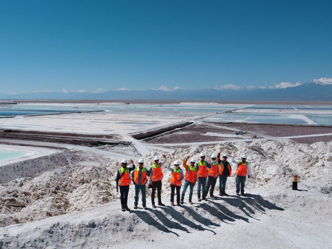 Group of people with safety helmets and bright orange safety vests standing on a hill in a salt mining area with large evaporation ponds; in the background, mountains and a clear blue sky are visible.