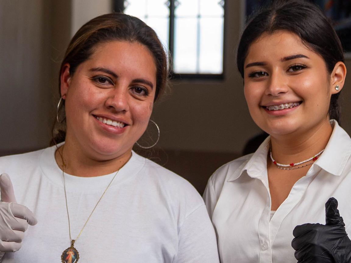 Drei Jugendliche in Arbeitskleidung mit Hörschutz strecken ihren rechten Daumen hoch und schauen in die Kamera. Vor ihnen stehen auf einem Arbeitstisch Holzprodukte wie gedrechselte Möbelbeine.
