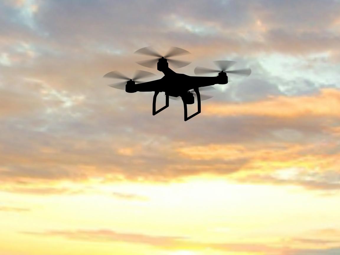 Die Silhouette einer Person, die eine schwebende Drohne per Fernsteuerung steuert, vor einem Abendhimmel