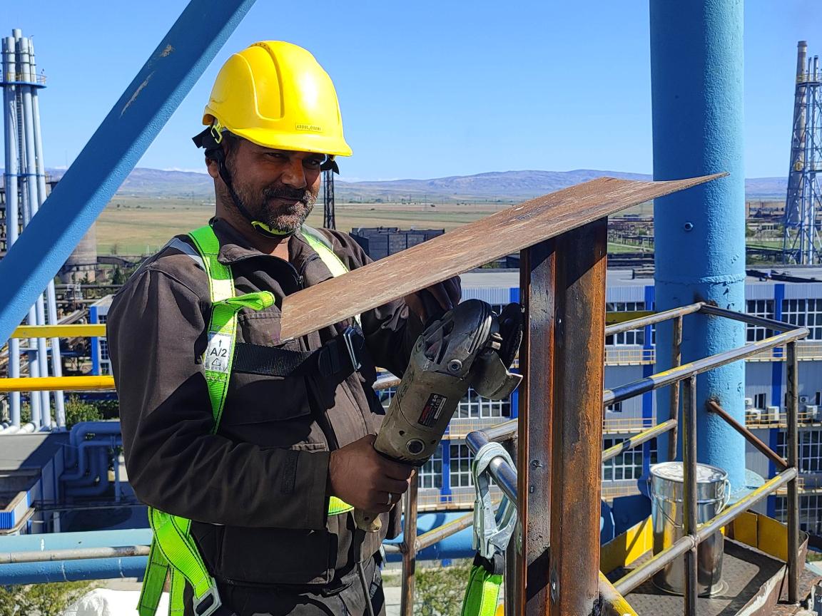 A man in a yellow helmet stands on a scaffold and grinds down a steel frame.