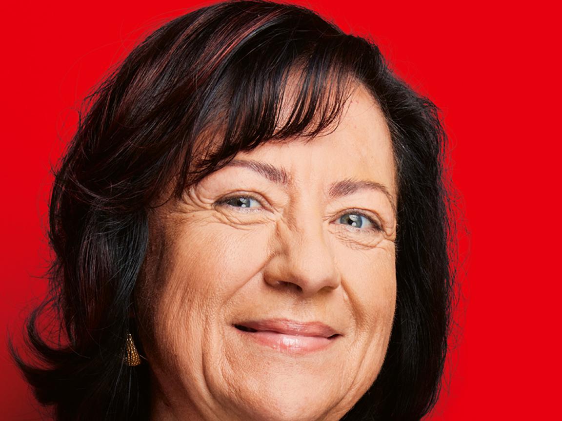 Porträt einer lächelnden älteren Frau mit dunklem, schulterlangem Haar und schwarzem Oberteil vor knallrotem Hintergrund.