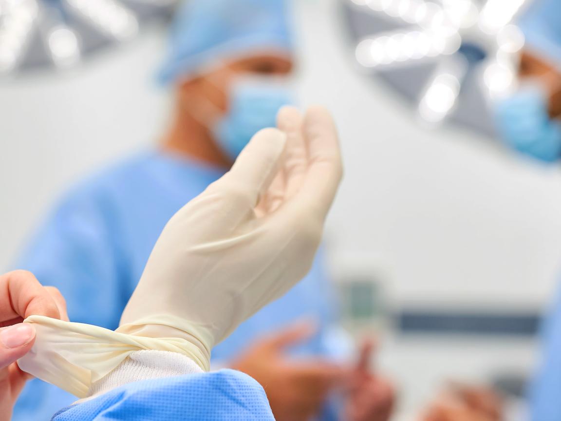 A medical professional is putting on sterile gloves in an operating room, with two other people in surgical attire blurred in the background.