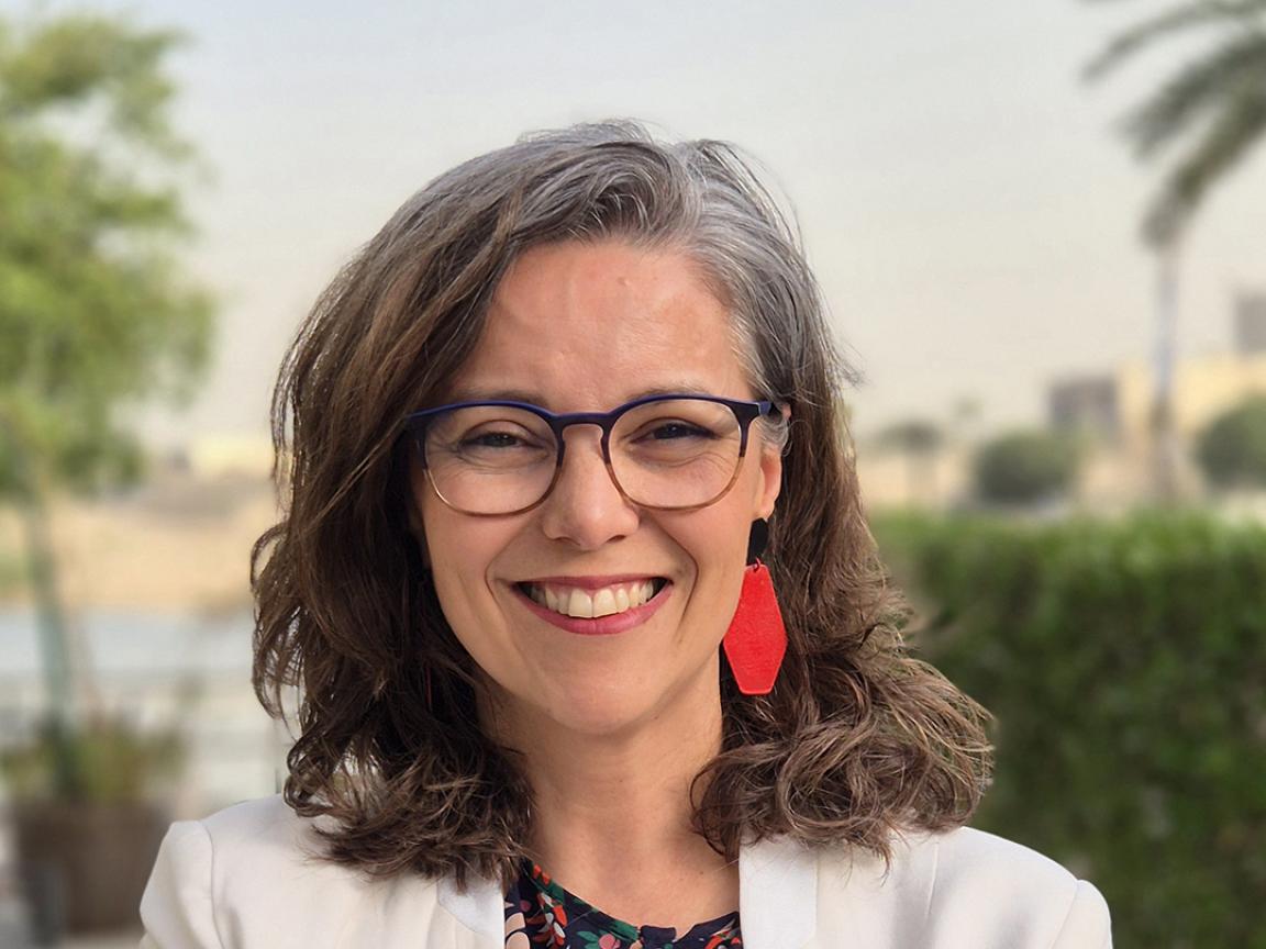 Porträt einer lächelnden Frau mit schulterlangem, braun-grauem Haar, rotem Ohrring, Brille und weißem Blazer vor einem unscharfen, grünen Außenhintergrund.