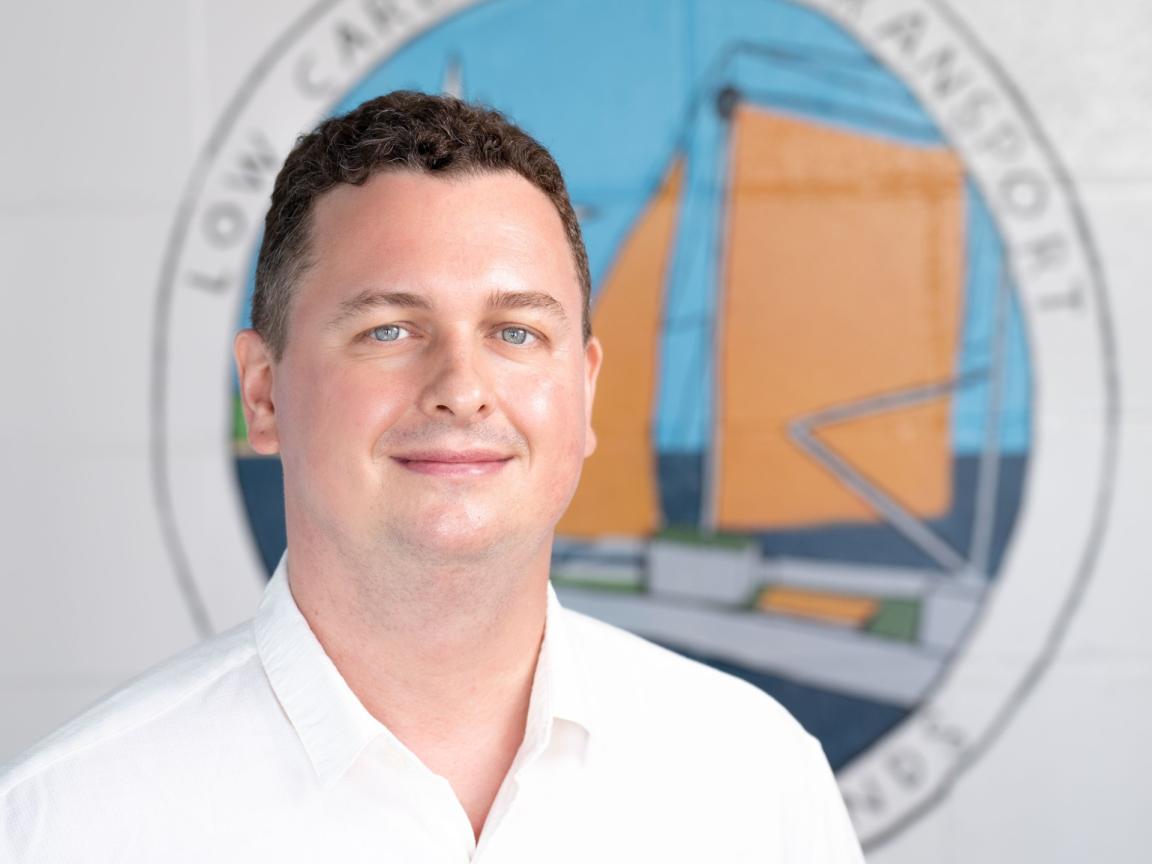 Porträt eines lächelnden Mannes mit kurzen braunen Haaren in weißem Hemd vor einem Wandbild mit der Aufschrift „Low Carbon Sea Transport“ und einer stilisierten Segelbootgrafik.