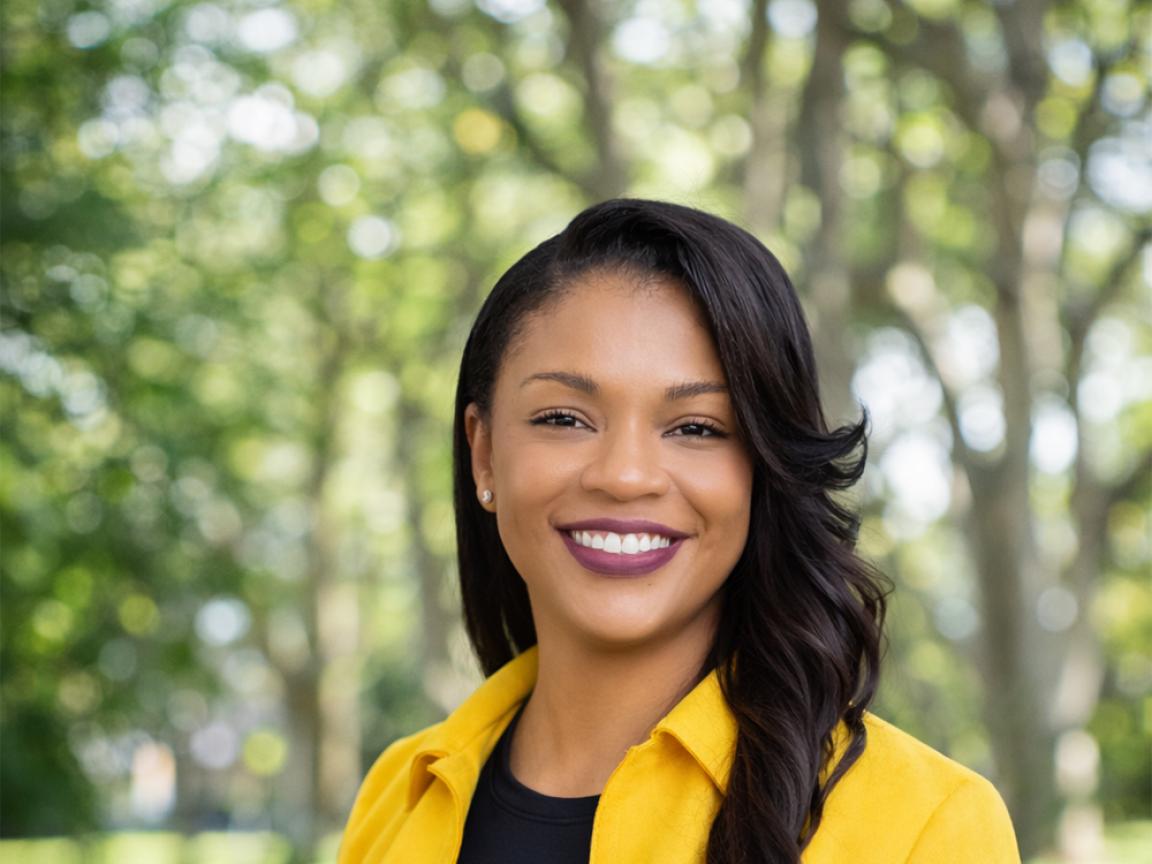 Portrait of a smiling woman with long dark hair wearing a yellow blazer and black top in front of a blurred green park background in daylight.