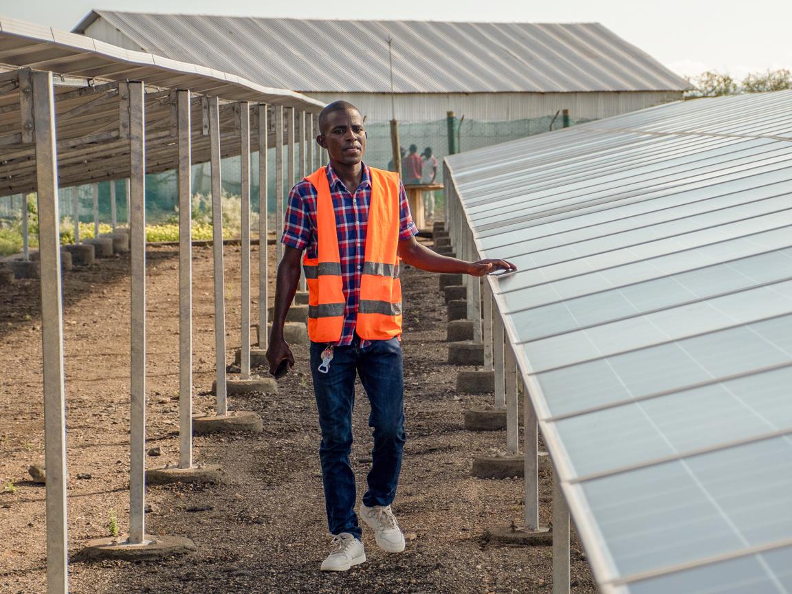 Man wearing an orange safety vest and checkered shirt standing next to a row of solar panels at an outdoor site with metal structures and buildings in the background.