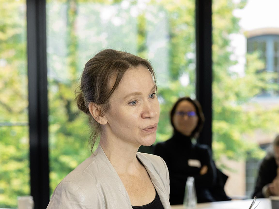 Frau mit zusammengebundenen Haaren hält ein Dokument in der Hand und spricht in einem hellen Seminarraum mit großen Fenstern, während im Hintergrund weitere Personen sitzen.