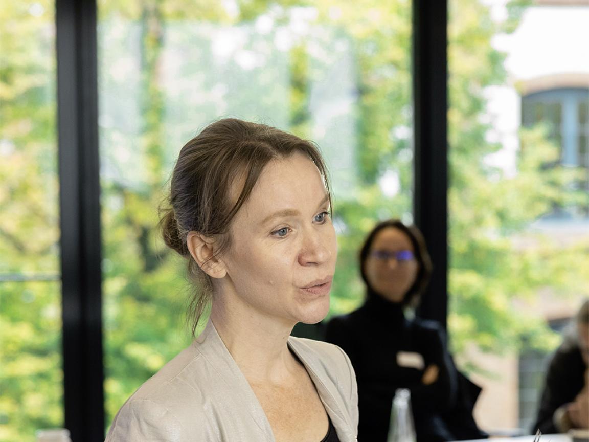 Frau mit zusammengebundenen Haaren hält ein Dokument in der Hand und spricht in einem hellen Seminarraum mit großen Fenstern, während im Hintergrund weitere Personen sitzen.