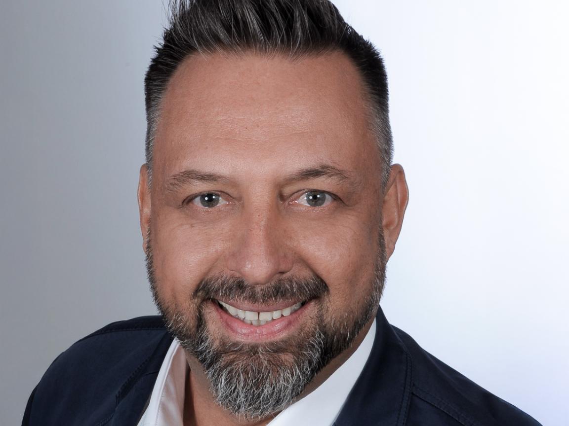 Porträt eines Mannes mit kurzem, grauem Haar und einem gepflegten Bart. Er trägt ein weißes Hemd und eine dunkle Jacke. Der Mann lächelt freundlich in die Kamera, vor einem neutralen, hellen Hintergrund.