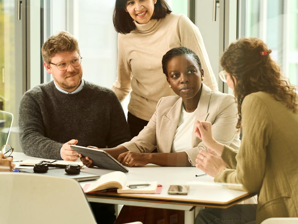 Several people sit at a table and discuss; a tablet and note-taking materials are on the table.