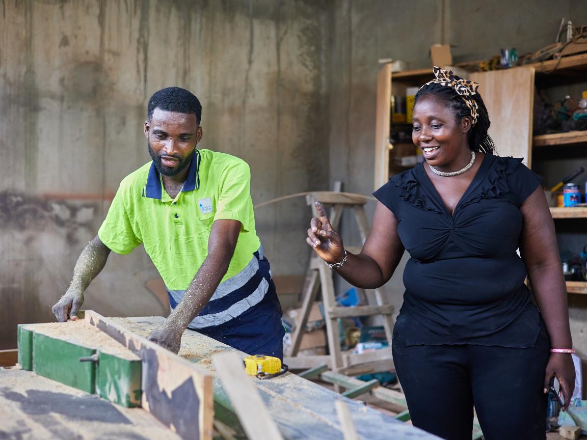 A man works at a woodworking machine; a woman stands smiling beside him, raising her hand