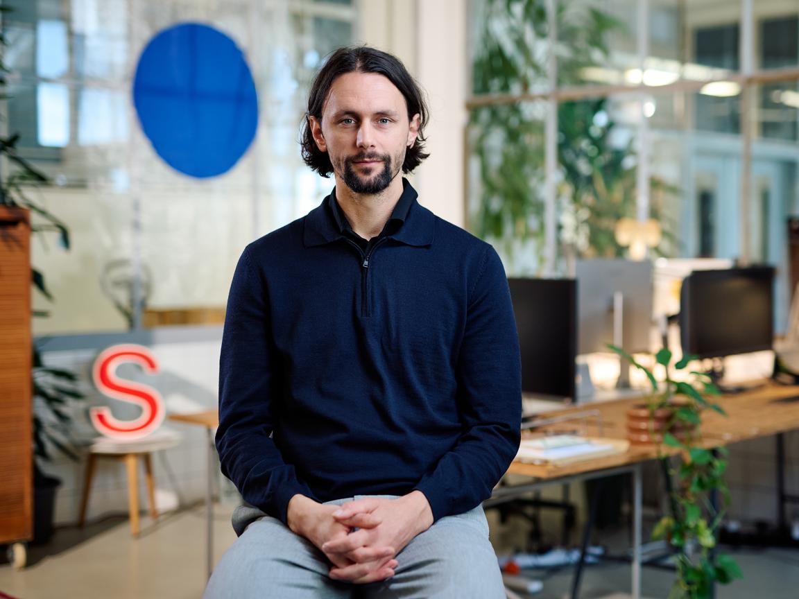 A man in a dark blue zip-up sweater sits with folded hands in a modern, plant-filled office, calmly looking at the camera.