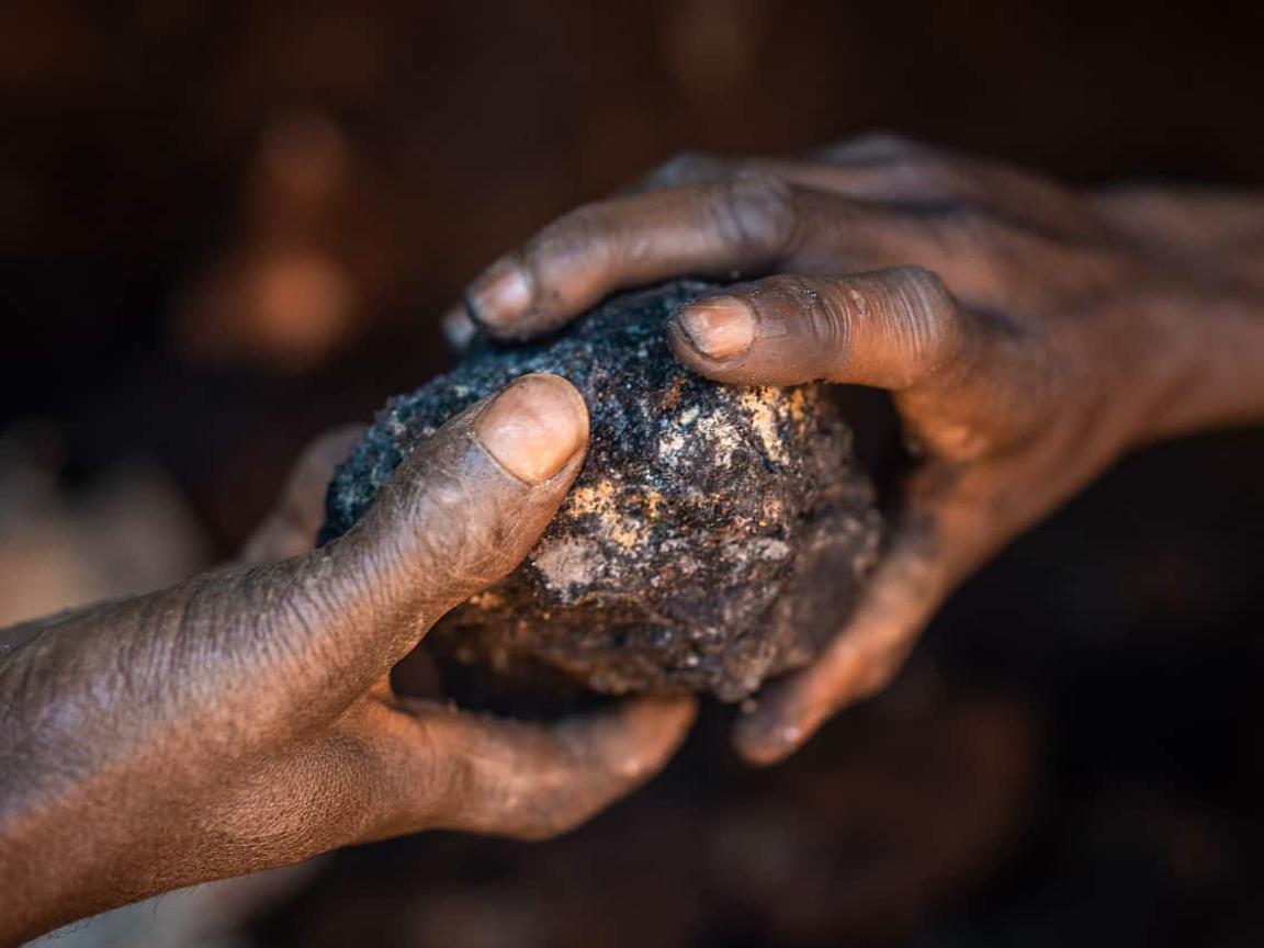 Zwei Hände halten einen dunklen, unbearbeiteten Gesteinsbrocken in einer schwach beleuchteten Umgebung.