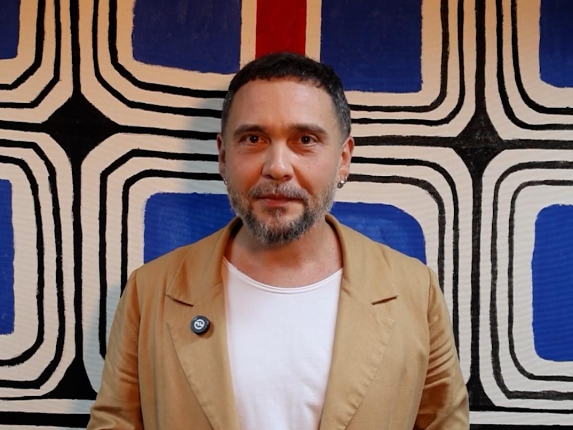 Ein Mann mit Bart in beigem Blazer und weißem T-Shirt spricht gestikulierend vor einem blau-schwarz gemusterten Wandgemälde.