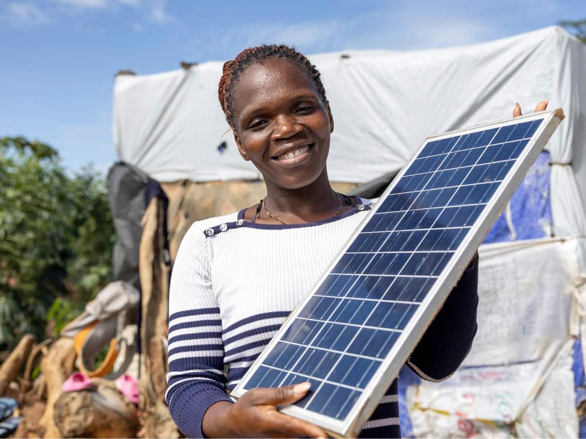 A woman smiles at the camera while holding a solar panel in front of a makeshift shelter.