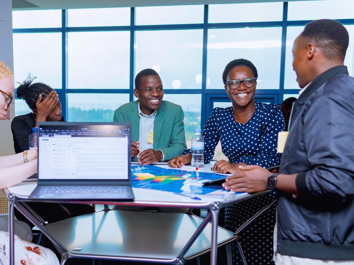 Mehrere Personen sitzen in einem hellen Raum an einem Tisch mit Laptop und Unterlagen und sprechen miteinander, im Hintergrund ein Banner „One Health Data Alliance Africa“.