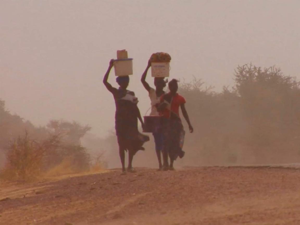Three people walk along a dusty road in haze, carrying containers on their heads.