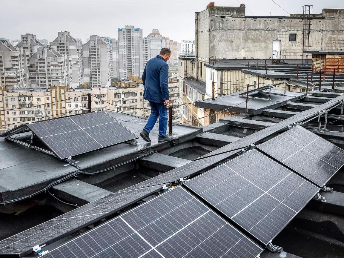 A man walks across a flat rooftop with installed solar panels, with many high-rise apartment buildings in the city behind him.