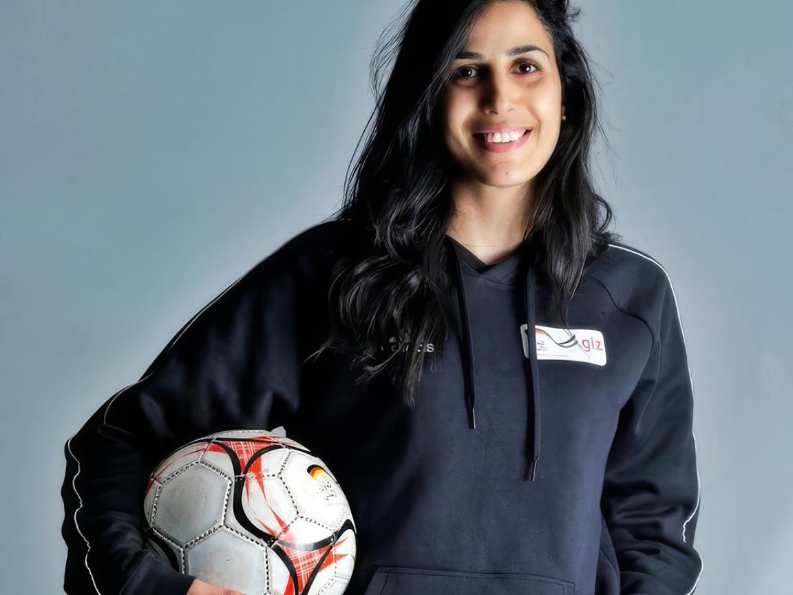 Porträt einer lächelnden Person mit dunklen langen Haaren, die vor grauem Hintergrund einen Fußball unter dem Arm hält.
