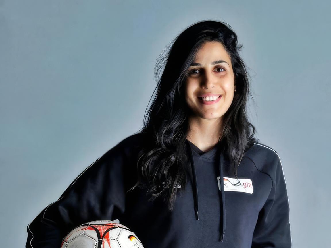 Portrait of a smiling person with long dark hair holding a soccer ball under one arm against a gray background.