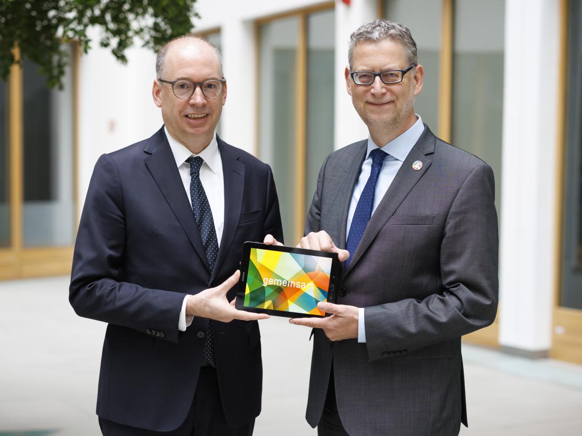 Zwei Männer in Anzügen und mit Krawatte halten gemeinsam ein Tablet mit bunter Grafik und dem Schriftzug Gemeinsam in den Händen
