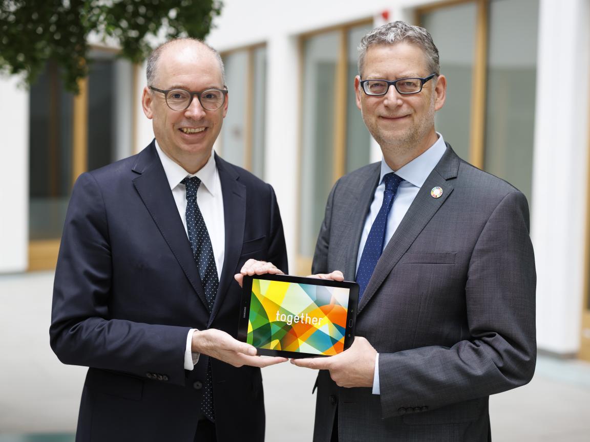 Two men in suits and ties are holding a tablet together, displaying colourful graphics and the word ‘Together’