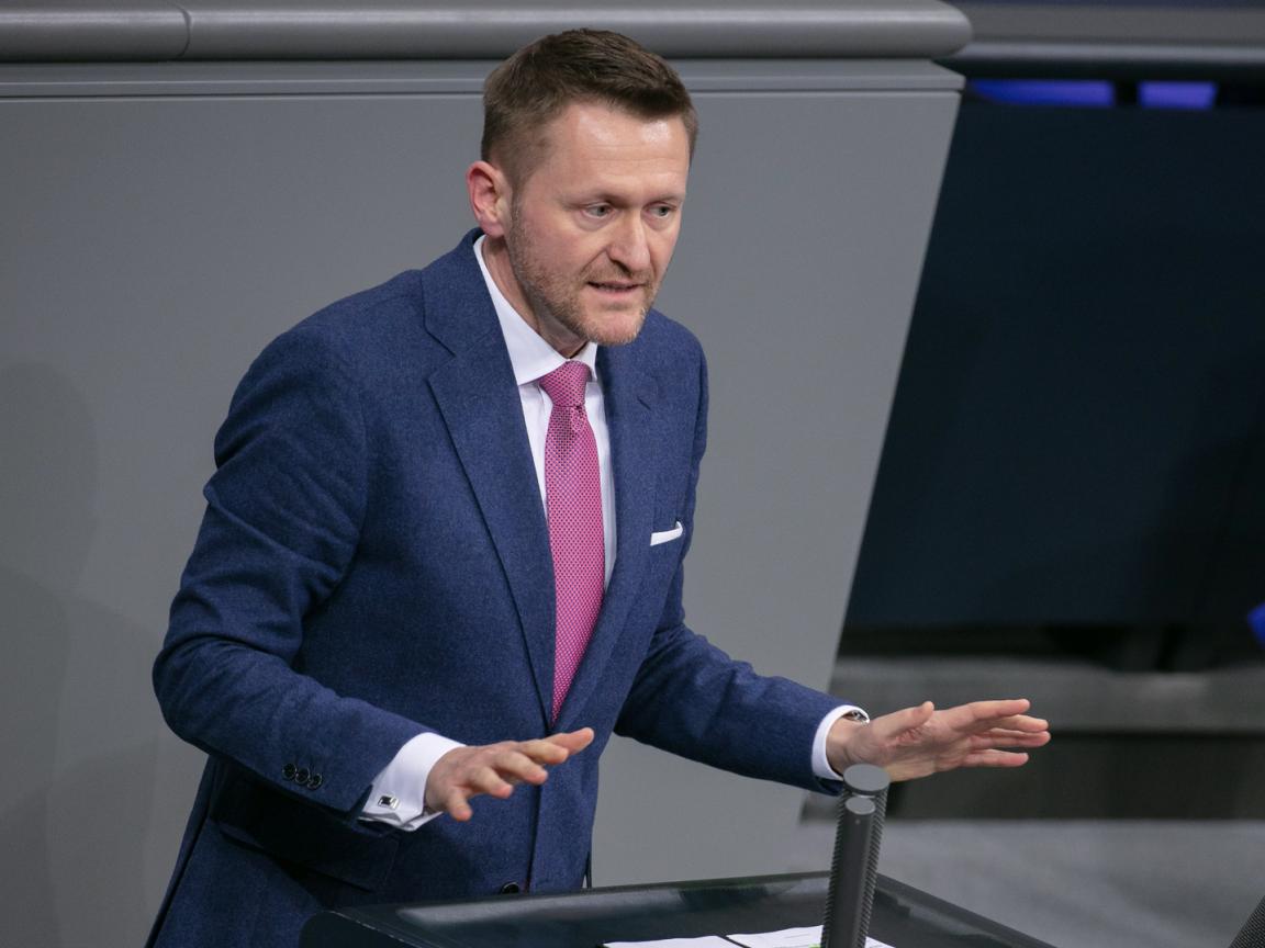 Ein Mann im blauen Anzug mit pinker Krawatte spricht gestikulierend an einem Rednerpult in einem Plenarsaal.