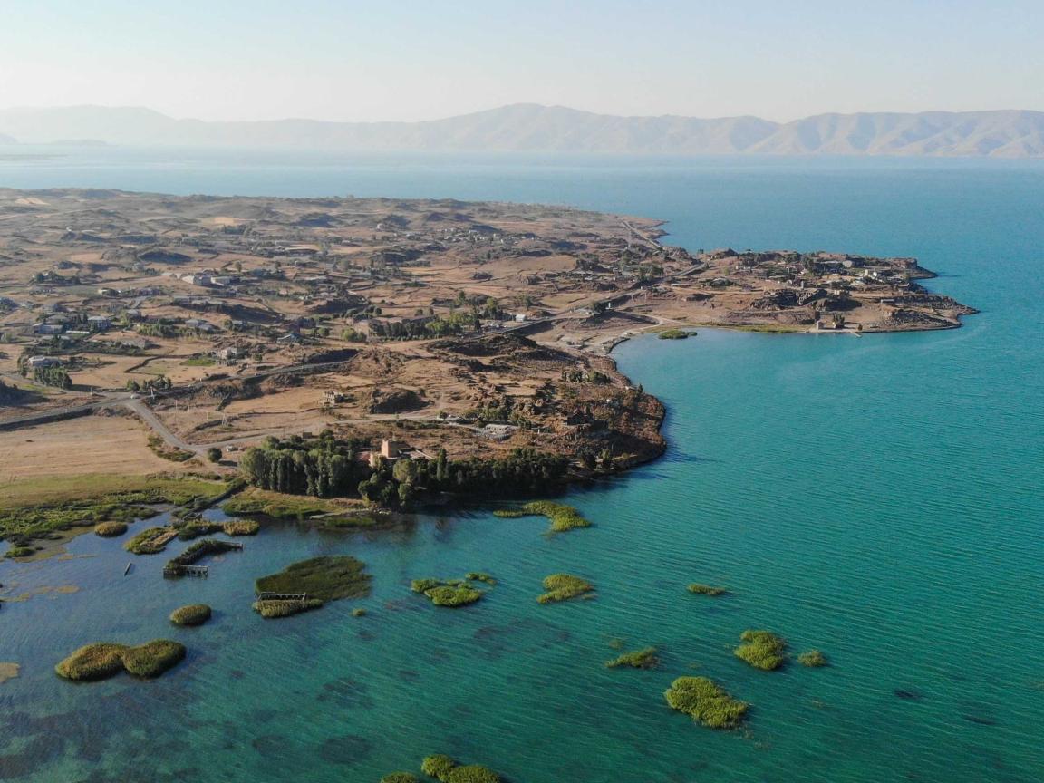 Luftaufnahme einer kargen Küstenlandschaft mit Bucht, türkisblauem Wasser und kleinen grünen Inselchen im Vordergrund sowie Bergen am Horizont.