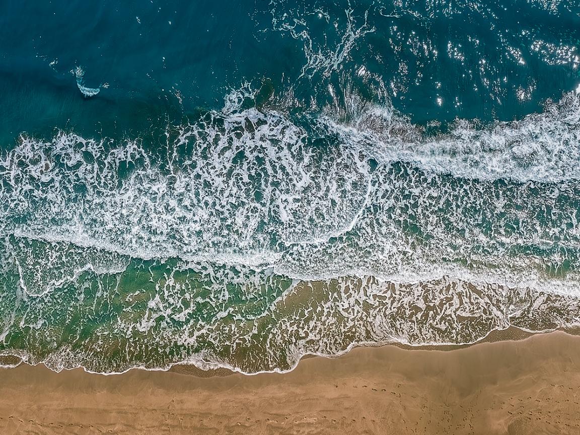 Luftaufnahme von Meereswellen, die mit weißer Gischt auf einen goldbraunen Sandstrand auflaufen, mit türkisfarbenem bis dunkelblauem Wasser im Hintergrund.