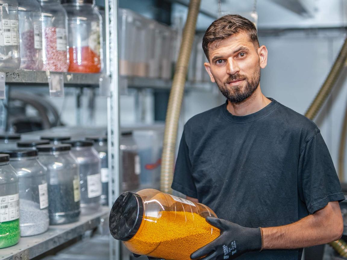 A bearded man in a dark T-shirt and black work gloves holds a jar of orange plastic granules and smiles at the camera, with shelves of containers filled with colored granules behind him.